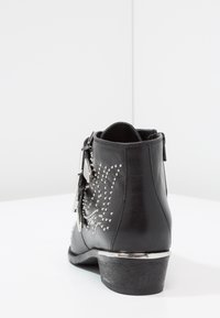 Botín negro con adornos de tachuelas, un acento en el tacón de plata brillante y una textura de cuero suave. Cierra con una cremallera en el lateral.