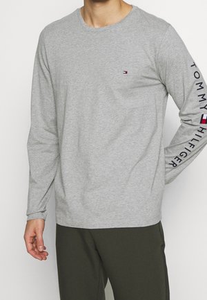 Mężczyzna w jasno szarej koszuli z długim rękawem, z małym logo z flagą na piersi oraz napisem "Tommy Hilfiger" na prawym rękawie.