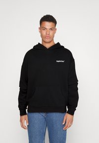 Svart hettegenser med frontlomme, har snor ved hetten og en liten hvit logo på brystet. Kombinert med blå denimjeans.