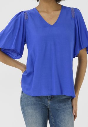 Femme portant un chemisier bleu à col en V avec des manches volantées et des accents en dentelle sur les épaules, associé à un jean en denim bleu.