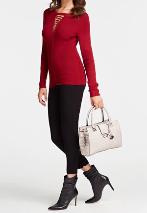 Femme portant un haut rouge à manches longues avec un détail à lacets sur le devant, un pantalon noir, des bottines noires et un sac à main blanc texturé.