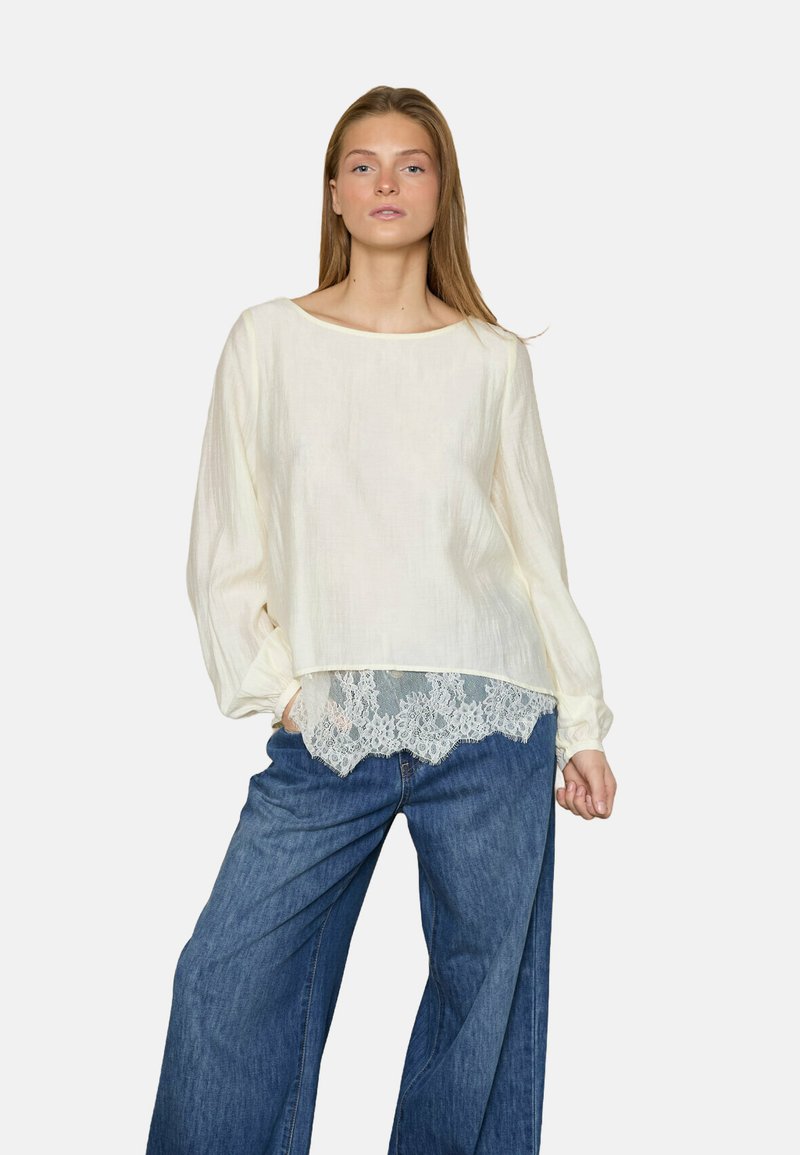 Cremefarvet bluse med brede ærmer og rund halsudskæring, præget af en blondekant i bunden. Bæres med løse blå denim bukser.