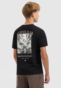 Tienerjongen die zijwaarts kijkt, draagt een zwart T-shirt met een geometrische stedelijke architectuurprint en de tekst "URBAN" op de rug, gecombineerd met bruine shorts.