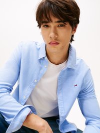 Camisa azul claro de botones con cuello, superpuesta a una camiseta blanca. Presenta un pequeño logo en el pecho y mangas enrolladas. Jeans de mezclilla visibles.
