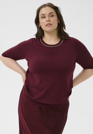 Burgunderfarbener Kurzarm-Pullover mit Rundhalsausschnitt, akzentuiert durch eine kettenartige Verzierung. Glattes Material, lockere Passform, kombiniert mit einem passenden Rock.