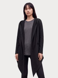 Kvinde med langt mørkt hår iført en sort asymmetrisk åben cardigan over en grå skjorte og sorte bukser mod en ensfarvet lys baggrund.