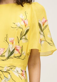 Robe jaune avec manches flottantes ornée de motifs de vignes florales roses et blanches et de feuilles vertes sur un tissu léger.