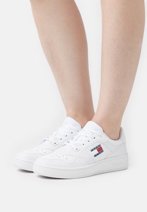 Hvide lavtop-sneakers med snøreføring og et Tommy Jeans-logo brugt på bare fødder mod en ensfarvet baggrund.