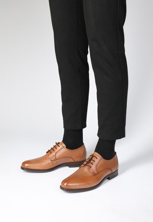 Brown leather dress shoes with a polished finish, lace-up design, rounded toe, and a low heel, paired with black ankle-length trousers and socks.