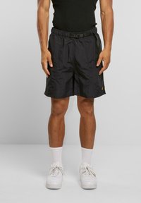 Svarte shorts med teksturfinish, elastisk midje med belte, sidelommer, og en liten logo på venstre bein. Kombinert med hvite sneakers.