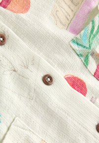 Light cream fabric with textured surface, featuring colorful fruit motifs. Includes wooden buttons and patch pockets for added detail.