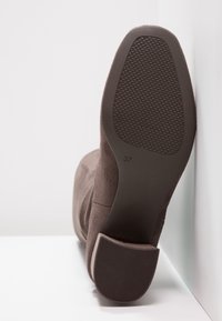 Bota de ante marrón con suela de goma texturizada. Presenta un tacón curvado y la marca de talla "37" en la parte inferior. Diseño minimalista.