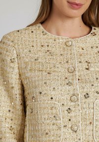 Beige strukturierte Jacke mit metallischen Pailletten, runden Knöpfen und weißen Paspelierungen an den Taschen. Kragenloses Design mit strukturierter Passform.