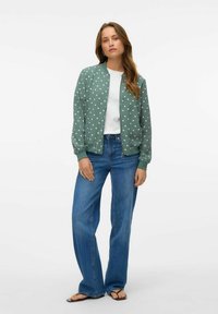Veste bomber verte à pois blancs, associée à un t-shirt blanc et un jean large bleu, portée avec des tongs noires.
