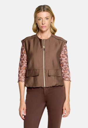 Femme aux cheveux blonds porte un gilet marron sans manches zippé sur un haut à manches longues à motifs et un pantalon marron assorti, debout sur un fond blanc.