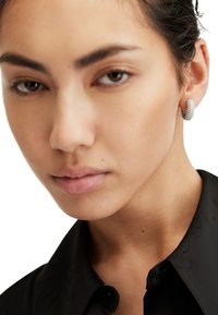 Boucles d'oreilles créoles argentées avec un design texturé en spirale. Gros plan sur un visage les portant, avec une peau lisse et des cheveux foncés. Chemise noire à col.