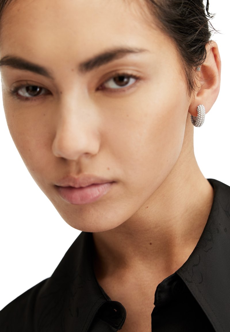 Boucles d'oreilles créoles argentées avec un design texturé en spirale. Gros plan sur un visage les portant, avec une peau lisse et des cheveux foncés. Chemise noire à col.