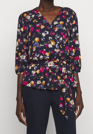 Vrouw draagt een marineblauwe wikkelblouse met roze, gele, witte en paarse bloemenprint, gecombineerd met effen donker marineblauwe broek.