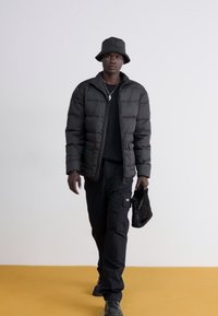 Giubbotto imbottito nero, maglietta nera a girocollo, pantaloni cargo neri, cappello a secchiello nero, con una borsa in ecopelliccia nera. Pavimento giallo.