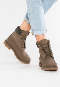 Bruine suède enkellaarsjes met ronde neus, vetersluiting, gevoerd kraagje en getextureerde rubberen zool. Draagt met opgerolde lichtblauwe jeans.
