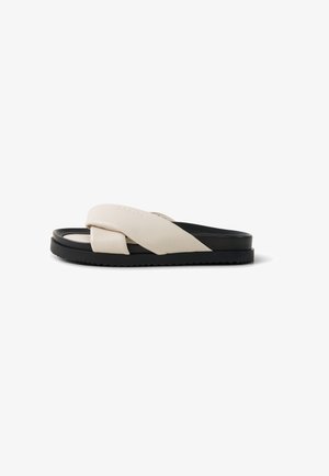 Sandale beige et noire à enfiler avec larges brides croisées rembourrées et semelle plate rainurée sur fond blanc.
