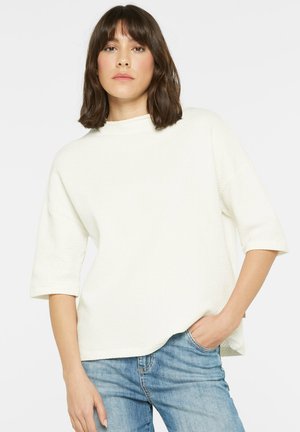 Frau mit schulterlangem braunem Haar, die ein lockeres weißes, strukturiertes Kurzarmoberteil und hellblaue Jeans trägt, eine Hand in der Tasche.