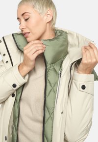 Witte donsjas met een groene gewatteerde voering, ritsopening en drukknoopsluitingen. De binnenkant toont een zachte, gestructureerde trui eronder.