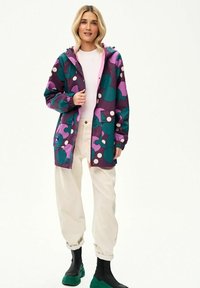 Kobieta o blond włosach, ubrana w fioletowo-zieloną wzorzystą kurtkę, białą koszulę, kremowe spodnie i czarne botki z zielonymi podeszwami, stojąca na białym tle.