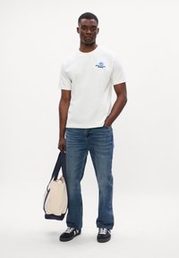 Valge lühikeste varrukatega T-särk sinise graafikaga, koos siniste teksade ja mustade tossudega. Hoides beežist ja mereväe sinisest käekotti.