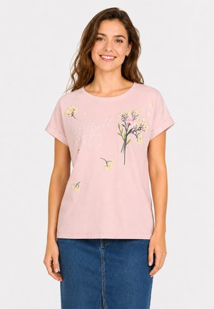 Young woman smiling, wearing a pink t-shirt with floral prints and "life full of joy" text, paired with a blue denim skirt.