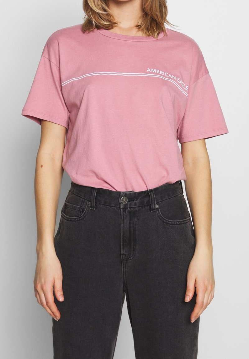 Personne portant un t-shirt rose American Eagle rentré dans un jean noir taille haute, debout devant un fond uni.