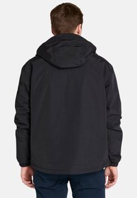 En man som bär en svart huvjacka, sedd bakifrån, med en neutral bakgrund. Jackan har elastiska muddar och en enkel design.