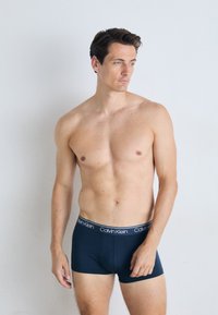 Mornarske boksarske hlače z elastičnim pasom Calvin Klein. Tkanina se zdi gladka in raztegljiva, kar poudarja tesno prileganje. Model stoji pokončno.