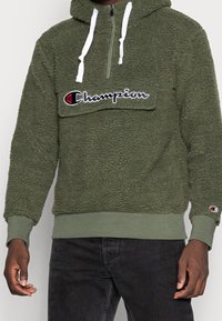 Felpa con cappuccio verde oliva in tessuto felpato, con zip a metà, tasca sul petto con logo ricamato, polsini e vita a costine. Tessuto textured dall'aspetto morbido.