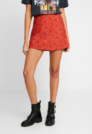 Vrouw draagt een rode minirok met bloemenprint, een zwart grafisch T-shirt en zwarte enkellaarsjes met gespen, staand tegen een effen achtergrond.