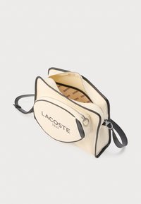 Kremfarvet lærredstaske med sorte detaljer, der har en rundet form og "LACOSTE 1933"-logo. Indeholder et lynlåslomme og justerbar rem.