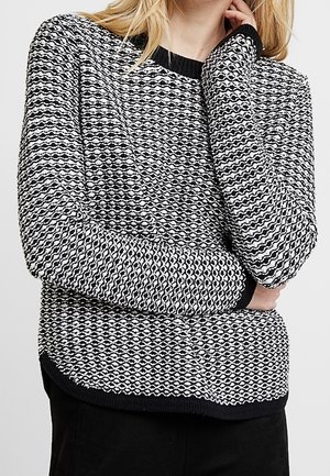 Femme portant un pull en maille texturée noir et blanc avec un motif ondulé, posant avec une main touchant son visage et l'autre bras croisé.