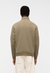 Mann mit dunkler Haut, der einen olivgrünen Pullover und hellbeige Hose trägt, steht mit dem Rücken zur Kamera vor einem schlichten weißen Hintergrund.