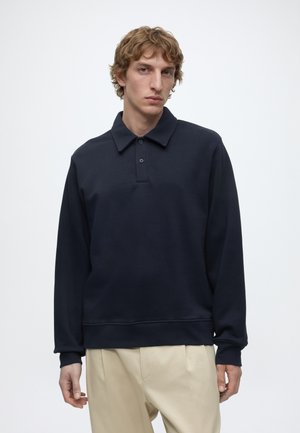 Młody mężczyzna o falowanych włosach, ubrany w ciemnogranatową koszulkę polo z długim rękawem i beżowe spodnie, stoi na jednolitym białym tle.