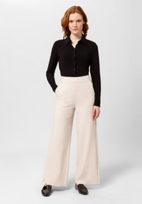 Černá košile s dlouhým rukávem a knoflíky, krémové kalhoty širokého střihu s vysokým pasem a tmavé boty. Hladká textura, elegantní střih a jednoduchý design.