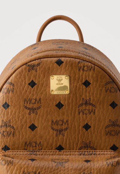 Καφέ δερμάτινο σακίδιο πλάτης MCM με μαύρο μοτίβο λογότυπου και ορθογώνια χρυσή πλακέτα με το σήμα της μάρκας που εμφανίζει τον σειριακό αριθμό στο μπροστινό μέρος.