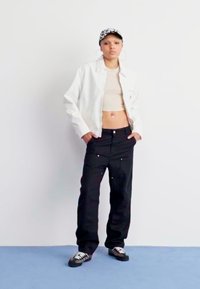 Chaqueta blanca corta sobre top beige, pantalones anchos negros y gorra con estampado. El calzado presenta un diseño en blanco y negro. Fondo liso.