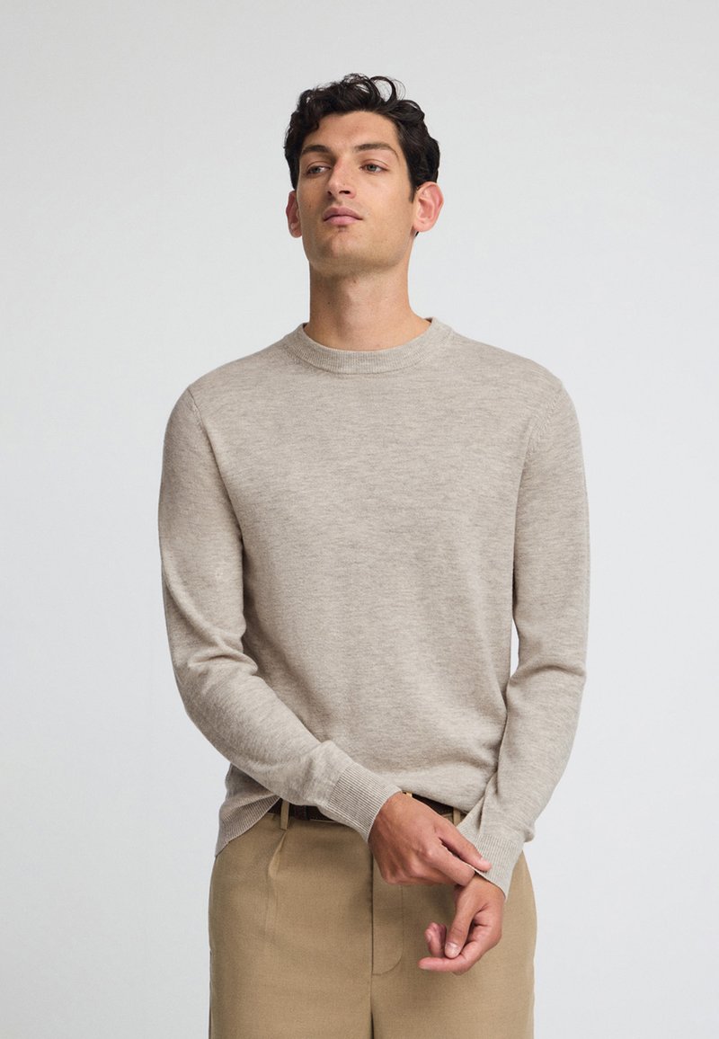 Beige langärmliger Strickpullover mit Rundhalsausschnitt und gerippten Bündchen, aus weichem Material, kombiniert mit beigen Hosen.