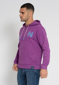 Päiväpeite, joka on valmistettu pehmeästä kankaasta, jossa on kenguruu-tasku, joka on varustettu erottuvalla sinisellä logolla rinnassa ja sinisillä kärjillä varustettu nyöri.