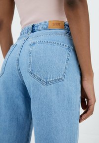 Henkilö, joka wearing vaaleansiniset farkut, joissa on takatasku ja nahkabrändipatch, yhdistettynä vaaleanpunaiseen hihattomaan toppiin.