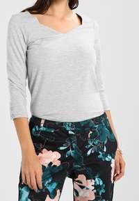 Graues Langarmshirt mit V-Ausschnitt und dekorativer Stickerei, kombiniert mit schwarzen Hosen im floralen Muster in Pink und Türkis.
