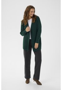 Skogsgrön, löst stickad cardigan med öppen front, bärs över en vit topp och svarta jeans. Djurtryckade klackstövlar fullbordar outfiten.