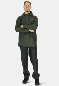 Donkergroene waterdichte jas met capuchon, drukknoopsluiting en zijzakken, gecombineerd met zwarte waterdichte broek en zwarte laarzen.