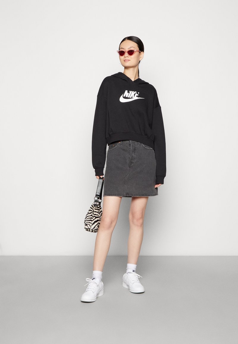 Sudadera recortada negra con el logo blanco de Nike, falda de mezclilla gris, bolso con estampado, zapatillas blancas y gafas de sol rojas en forma de gato.