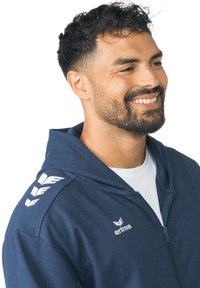 Marineblauwe hoodie met een ritssluiting, zijzakken en witte merkdetails op de mouwen en borst, gemaakt van zachte, gestructureerde stof.